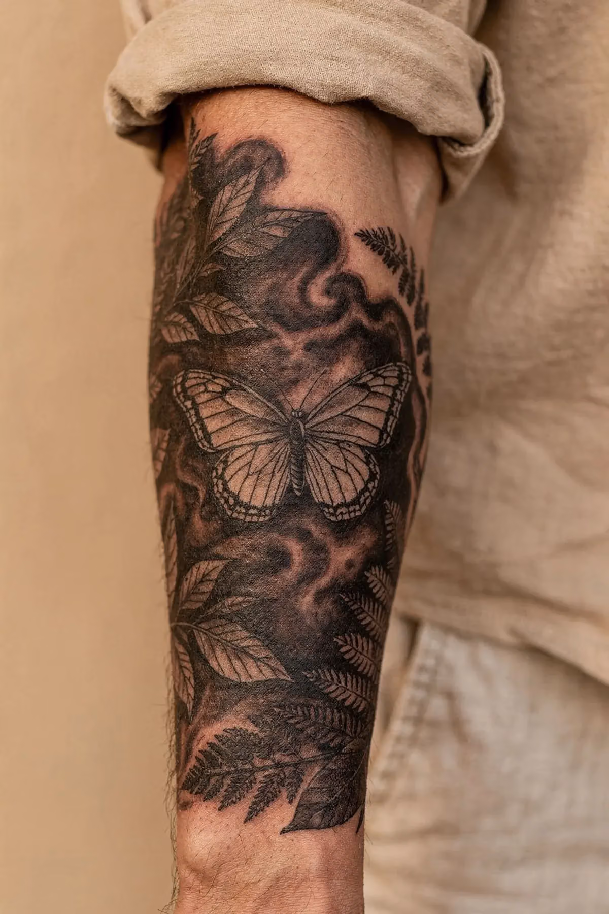 Schmetterling Tattoo in hellem Grau als Mittelpunkt eines dunklen Halbarm-Sleeves aus Rauch und Farnen