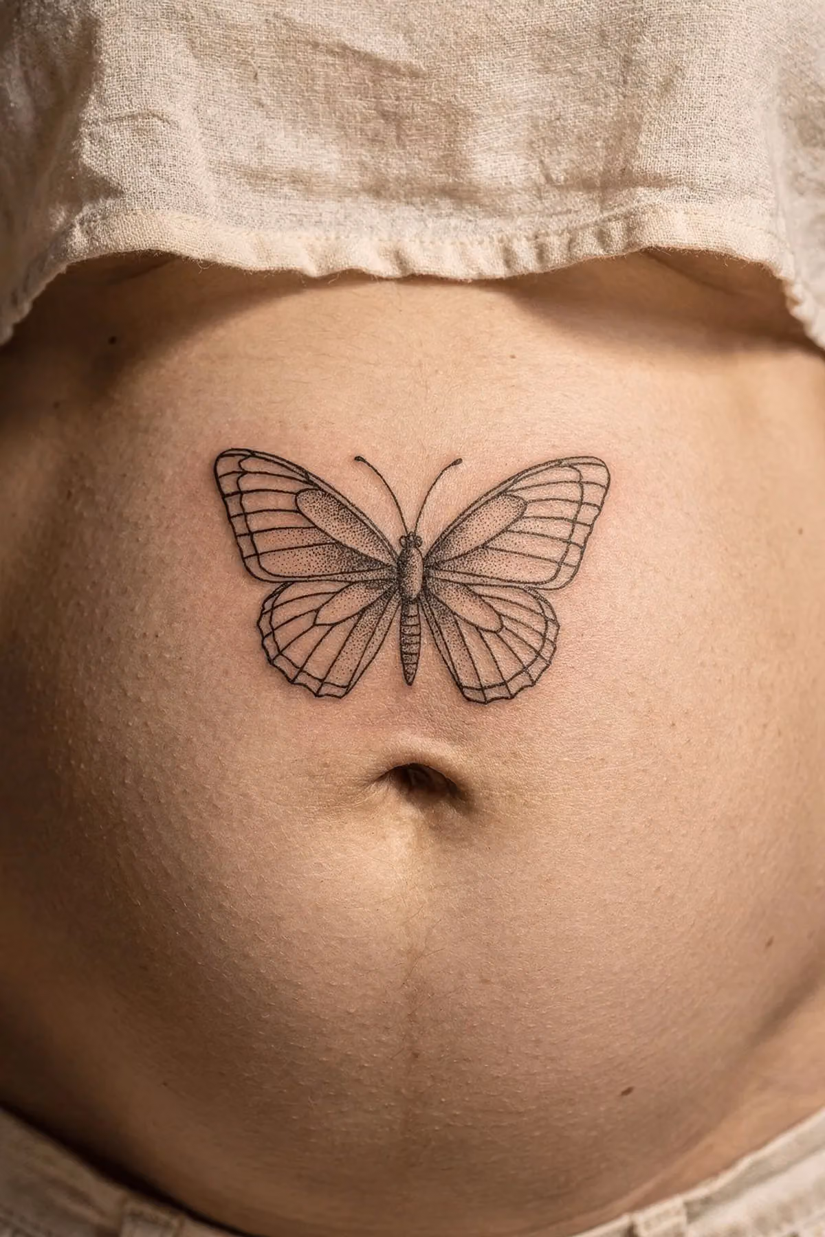Nahaufnahme eines Bauchs mit einem feinen schwarzen Schmetterling-Tattoo direkt über dem Nabel.