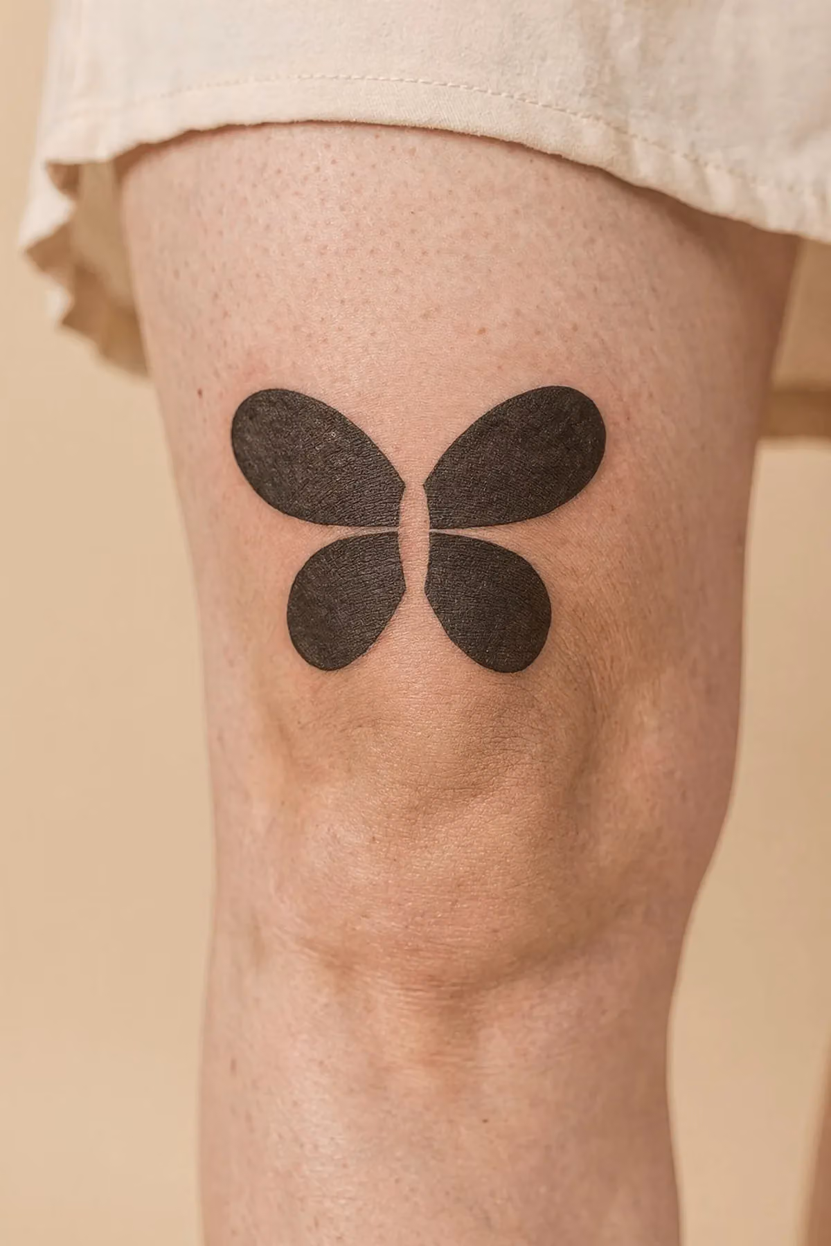 Schwarzes Tattoo auf gebeugtem Knie, das durch eine geteilte Ovalform einen fliegenden Schmetterling bildet.