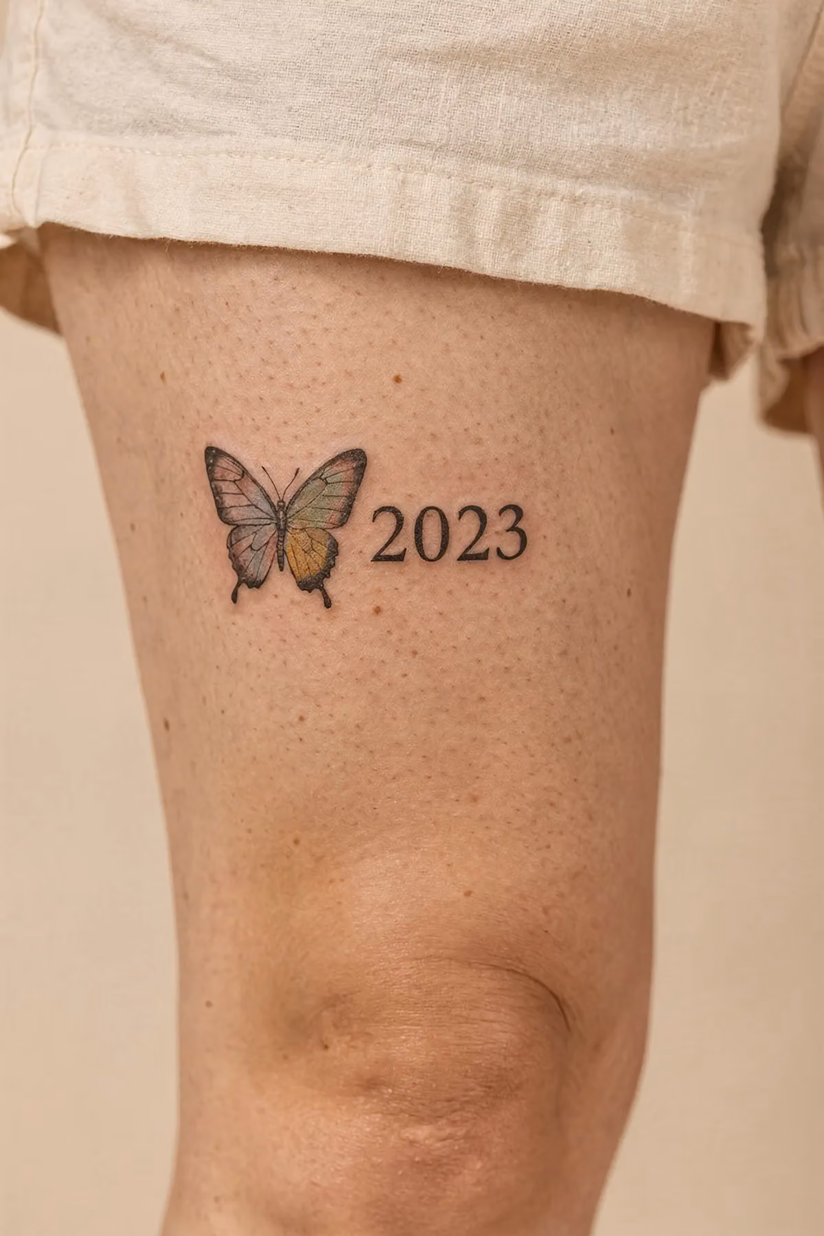 Kleiner Schmetterling mit pastellfarbener Flügelakzent und Jahreszahl 2023 am Oberschenkel