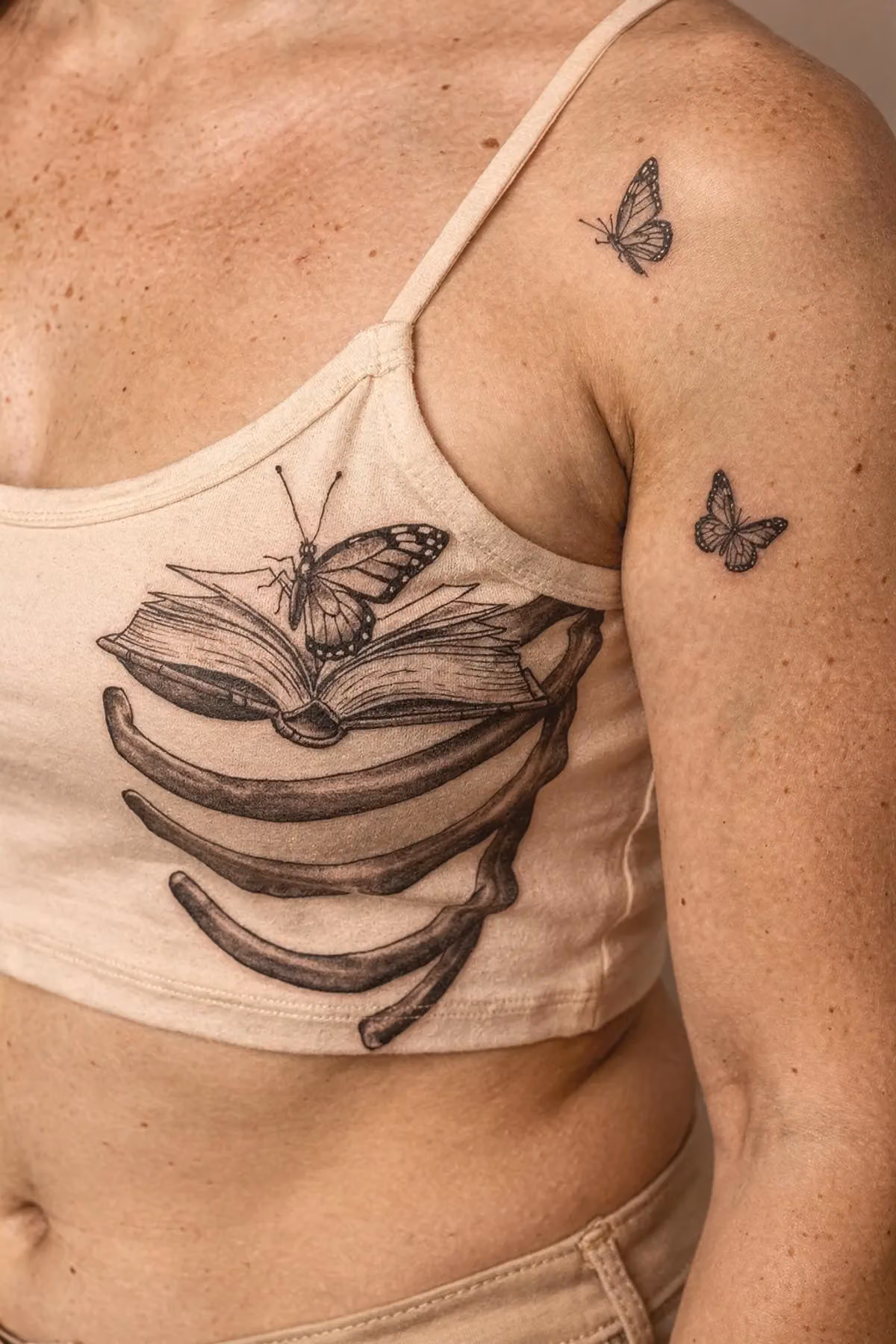 Vordere Schulter nahe Schlüsselbein mit zartem Schmetterlingstattoo und weißen Akzenten neben Trägerkante