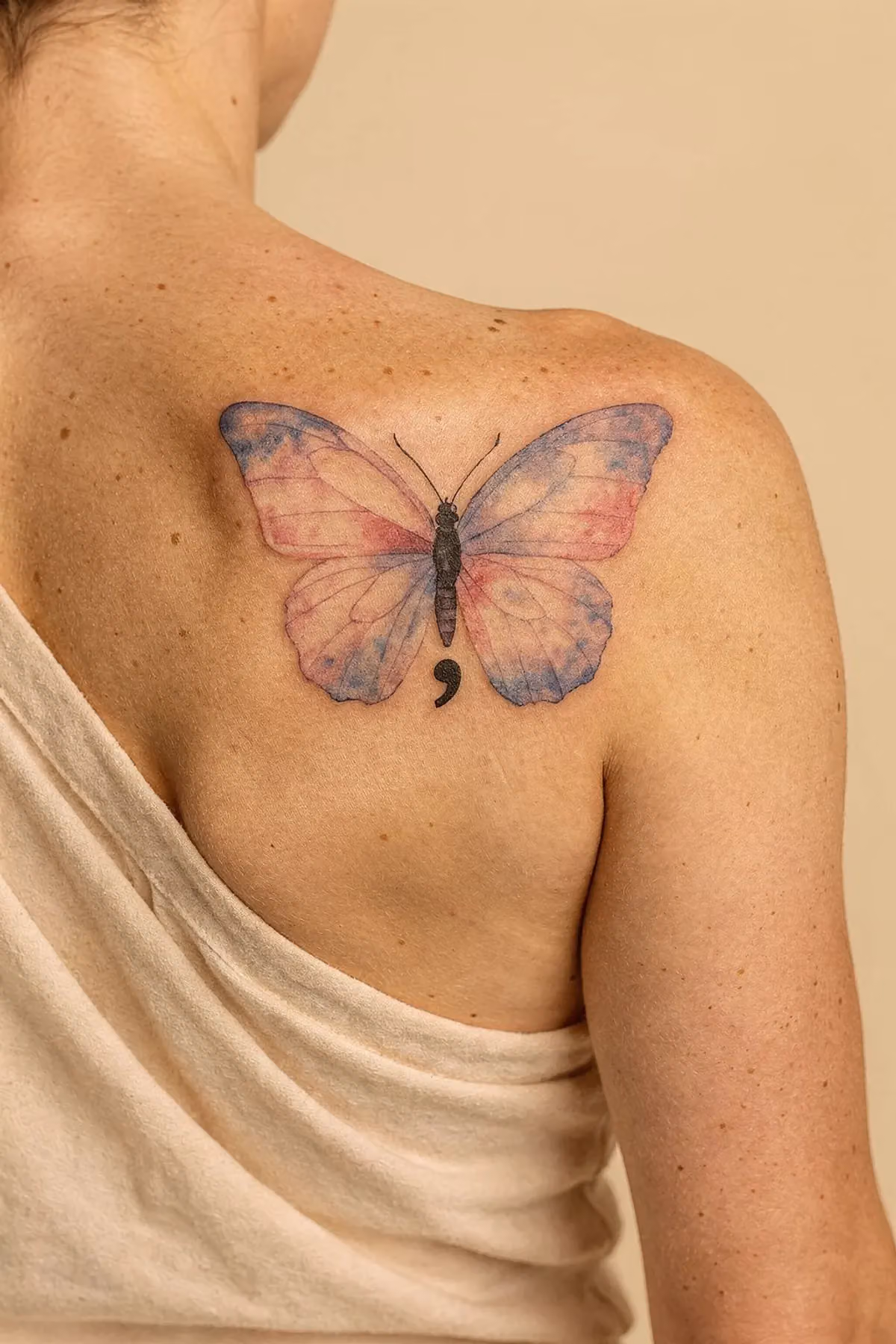 Schulterblatt mit einem Aquarell-Schmetterling: transparente Pastellverläufe in Rosa, Lavendel und Blau, das Semikolon tiefschwarz
