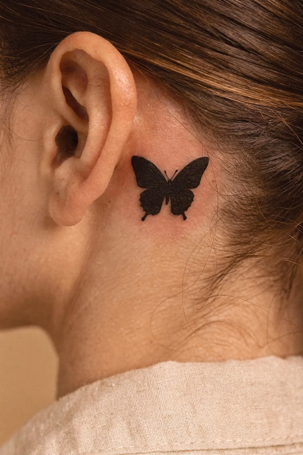 Kräftig schwarz ausgearbeiteter kleiner Schmetterling auf der Haut hinter dem Ohr.