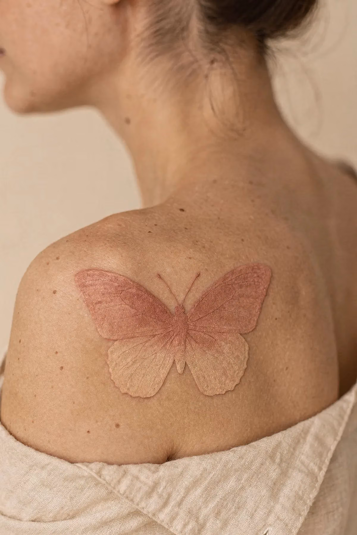 Schmetterling auf der Schulter, dessen Flügel als weicher Verlauf von Altrosa zu blassem Pfirsich ohne Umrisslinie gestochen sind.